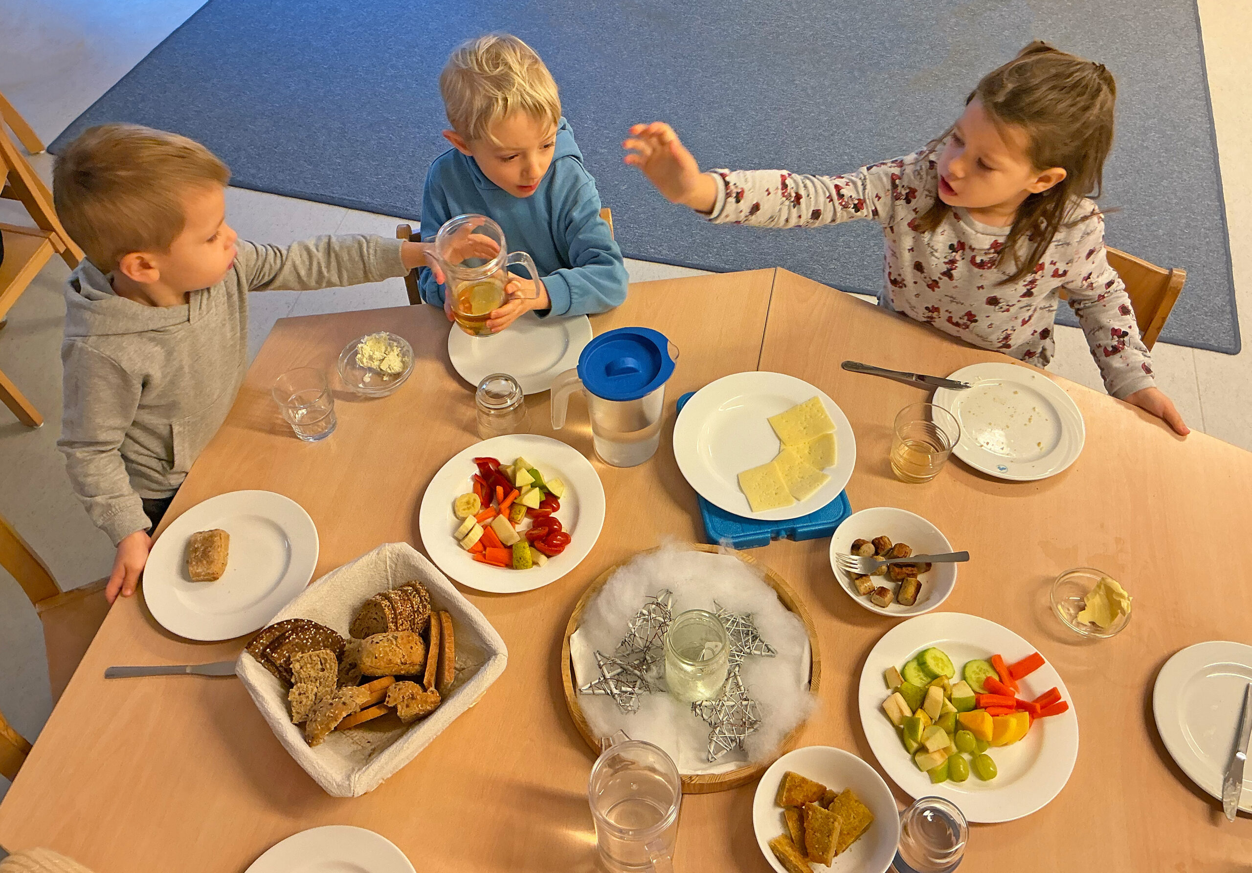 Drei Kinder sitzen in der Kita am Frühstückstisch und reichen einen Getränkekrug rum